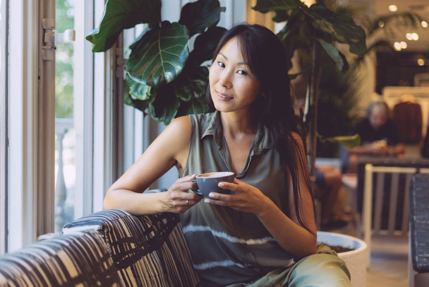 魅力的なアジア学生の長い黒髪の女性は、居心地の良いカフェに座りながら朝のコーヒーを楽しんでいます。一杯のカフェでコーヒーと座っているカジュアルな服を着ている内気な少女の肖像画. - 写真・画像