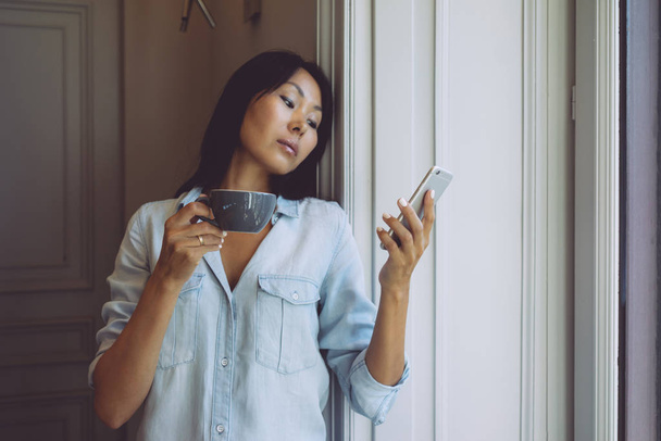 Attractive asian woman is surfing the web by a mobile phone while having break with cup of coffee in modern coffee shop. Modern designer is reading emails on a smartphone connected to wi-fi - Fotó, kép