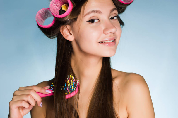 a smiling young girl makes a fashionable hairstyle, on her head a big pink curler, combs her hair - Foto, Imagen