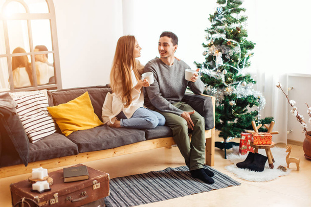 Man and woman hold cups of hot drink sitting before white Christmas tree - Foto, Imagem