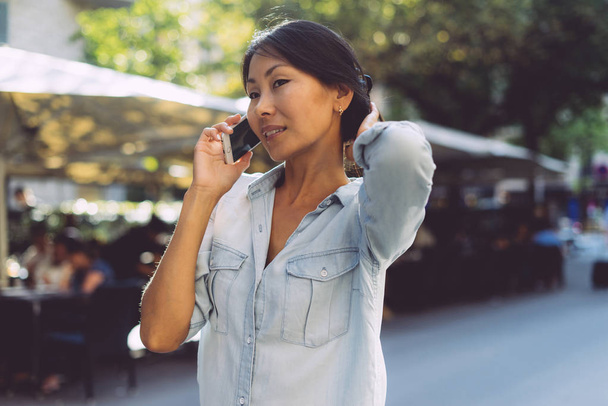 Beautiful asian woman with dark hair wearing jean dress is talking by a mobile phone while standing on a blurred street background. International student is chatting with friends by a smartphone. - Foto, imagen