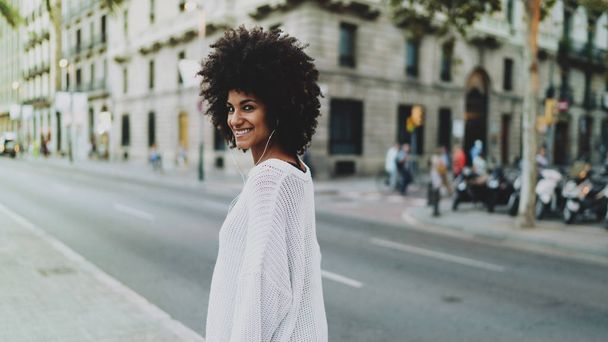 Αφρο αμερικανικό γυναίκα να ακούτε μουσική στα ακουστικά ενώ το περπάτημα στην πόλη με το κινητό τηλέφωνο. Freelancer θηλυκό χαλάρωση στους εξωτερικούς χώρους με μια μουσική κατέβασα και έπαιξε σε ένα smartphone. - Φωτογραφία, εικόνα