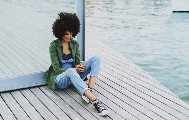 Smiling attractive student female with afro hairstyle wearing jeans overalls is typing messages on a display of a mobile phone while sitting on a bench at the city berth area on a day. - Фото, изображение