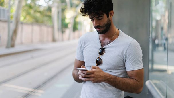 Professional web designer reading messages from colleagues working on a weekend on a new project. Young bearded guy using transport application on a mobile phone while waiting for the bus. - 写真・画像