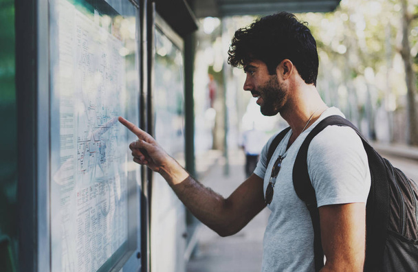 Young bearded traveler man searching the route on a public transportation system map in unknown city. Handsome guy with backpack pointing finger on train timetable on a railway station. - Foto, Imagen