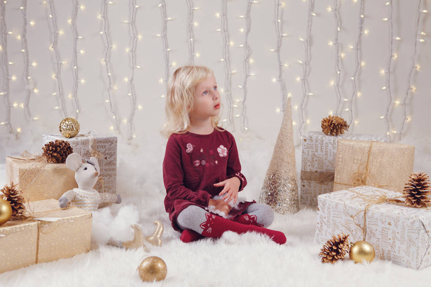 Porträt eines weißen blonden kaukasischen Mädchens mit blauen Augen, das auf dem Boden sitzt und mit Spielzeug Weihnachten oder Neujahr feiert. - Foto, Bild