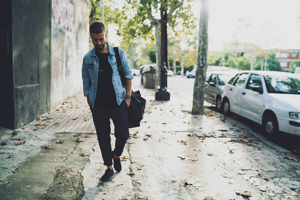 Doordachte knappe bebaarde man in spijkerbroek jas lopen de straat van de stad op een zonnige herfstdag. Stijlvolle jonge internationale student buiten wandelen met een zak op zijn schouder op een dag sweekend. - Foto, afbeelding