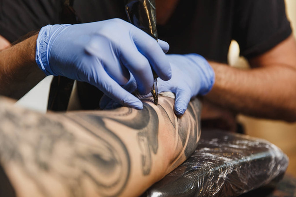 Közelről szakmai tattooer művész csinál kép a kezét, az ember a gép fekete tinta a jar. A test tetoválás minták. A tetoválás művészet berendezés. Mester teszi tetovált világos stúdió. - Fotó, kép