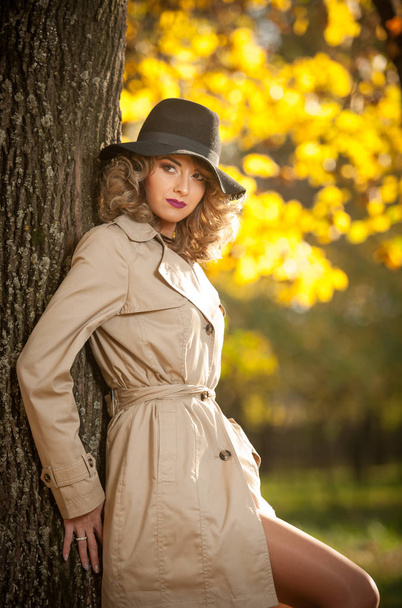Beautiful blonde woman with cream coat, long legs and black hat in a autumn scene.  Portrait of a very beautiful, elegant and sensual woman with curly hair and sexy legs  posing in autumn park. - Fotografie, Obrázek