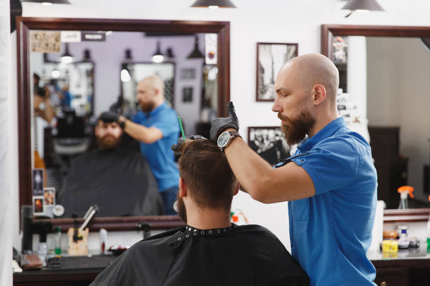 Мужчина профессиональный парикмахер, обслуживающий клиента клиппером. Рыжий красивый грубый стильный молодой человек с густой бородой и короткими волосами, стригущийся в черном плаще. Светло-белая парикмахерская
 - Фото, изображение