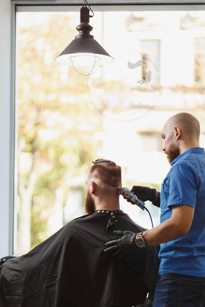 Male professional hairdresser serving client by clipper. Ginger handsome brutal stylish young man with thick big beard and short hair getting trendy haircut in black cape. Light white barber shop room - Photo, Image