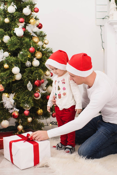 Щасливий молодий батько з милим маленьким сином у червоній шапці. Дитячий хлопчик сидить у світлій кімнаті вдома з прикрашеною новорічною ялинкою та подарунковими коробками. Різдвяний гарний настрій. Концепція сім'ї, любові та свята 2018
. - Фото, зображення
