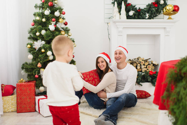 Happy young cheerful parents with cute little son in red hat. Child boy in light room at home with decorated New Year tree and gift boxes. Christmas good mood. Family, love and holiday 2018 concept. - Zdjęcie, obraz