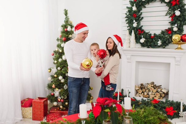 Happy cheerful parents with cute little son. Child boy with big red tree toy ball standing in decorated New Year light room at home on the background of Christmas wreath. Family, holiday 2018 concept. - Photo, Image