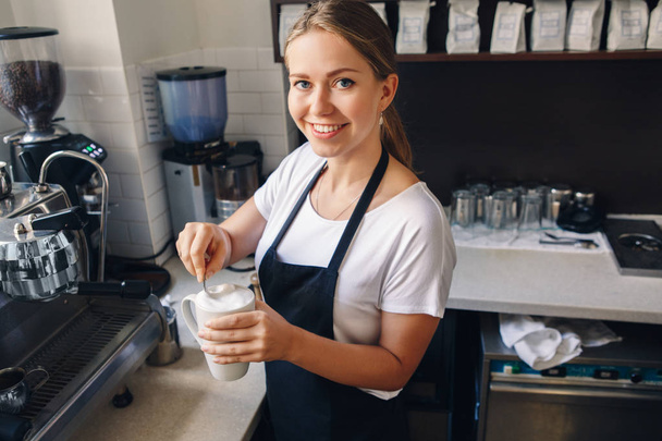 Portrét mladé krásné Kavkazský žena barista míchání pěny v šálku kávy cappuccino. Člověk na práci, malé obchodní koncept - Fotografie, Obrázek