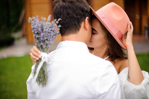 Groom in white shirt hugging bride in white dress and pink hat with bouquet of flowers - 写真・画像