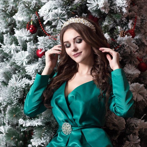 A beautiful girl with long hair in a green dress with the white diadem on her head near the Christmas tree . A square picture. - Photo, image