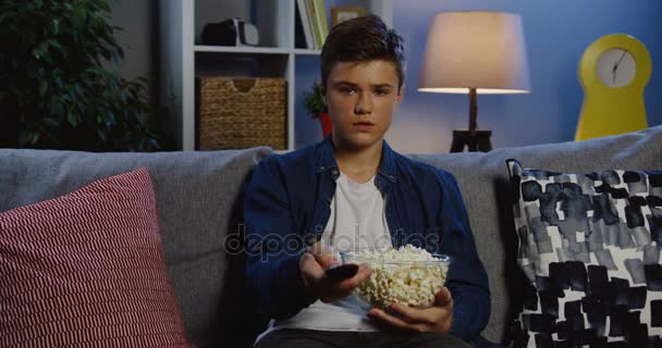 Teenager with a popcorn changing channels and choosing what to watch on TV in the nice living room in the evening. Indoor. Portrait shot - Metraje, vídeo
