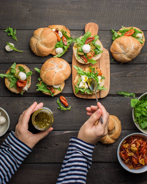 Sandwiches with arugula, dried homemade tomatoes, pesto sauce, mozzarella. Cooking process. Food photo with hands. Atmospheric food photo with wooden dark background. Place for text. Flat lay, top view - Photo, Image