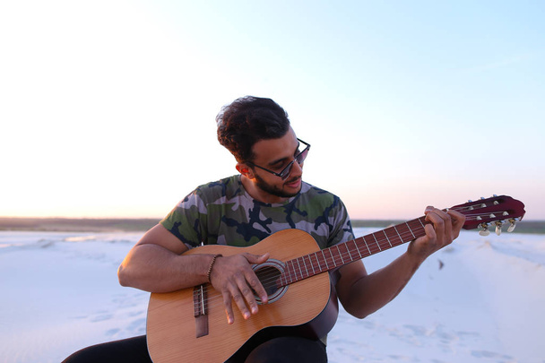 Аравійська гарний юнак налаштовує гітара, сидячи на пагорбі в мі - Фото, зображення