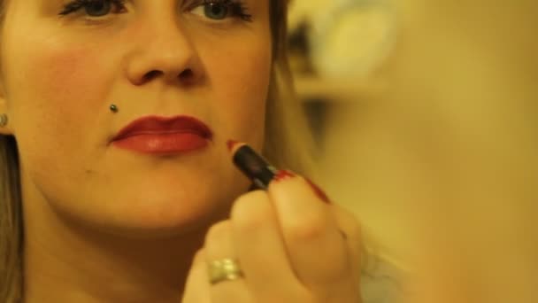 Young beautiful blond woman paints her lips bright red color lipstick in front of a mirror - Materiał filmowy, wideo