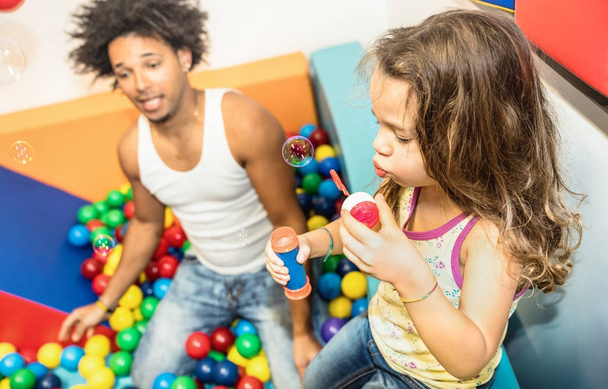 Latin american dad playing with mixed race daughter on ball pit pool at kindergarten playroom - Family concept with happy multiracial child and father having fun at kid toyroom with soap bubble blower - Foto, imagen
