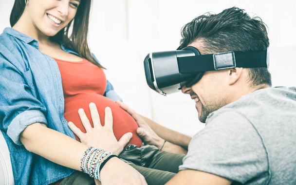Fiatal pár szórakozik a vr szemüveg tech fejhallgató - boldog ember ember férjével, virtuális valóság eszköz segítségével a felesége hasa - hordható technológia koncepció - tiszta vintage szűrő terhes nő - Fotó, kép