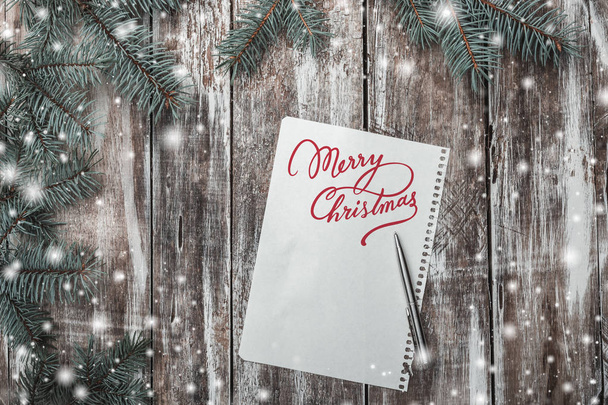 メリー クリスマスのメッセージをそれの上に白いシートで古い木製の背景。冬の休日にあたってお祝いメッセージのためのスペース。左右の側面を緑のモミの枝。雪の効果。平面図です。水平方向に. - 写真・画像
