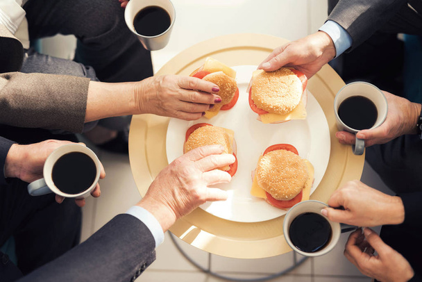 Close-up photo of four hamburgers on a plate and four hands. People are holding cups of coffee. - Fotó, kép