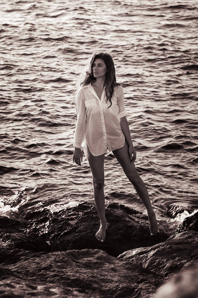 Musta ja valkoinen kuva nuoresta naisesta, jolla on pitkät ruskeat hiukset, pitkät hoikat jalat yllään valkoinen paita. Nainen seisoo kivien äärellä vedessä
 - Valokuva, kuva
