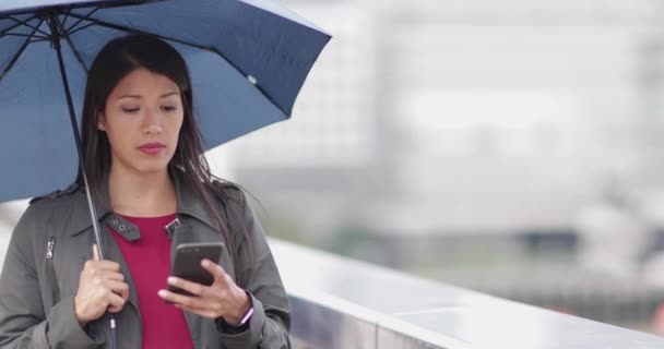 Businesswoman in city holding umbrella looking at smartphone - Imágenes, Vídeo