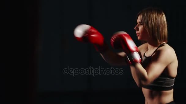 Morena en traje deportivo y guantes de boxeo sobre un fondo negro conduce golpes de entrenamiento en el aire. Boxeo de sombras
 - Imágenes, Vídeo