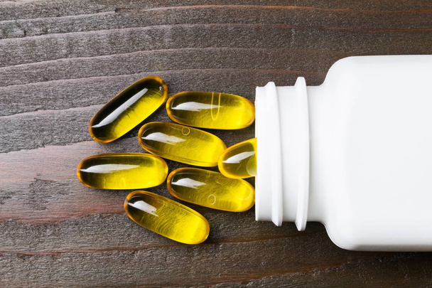 Cápsulas amarillas omega 3, ácido graso, aceite de pescado con botella blanca sobre mesa de madera oscura, macro imagen, vista superior
. - Foto, Imagen