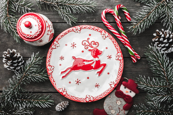 Christmas greeting card on wooden background, fir branches, cones, a snowman, dishes with Christmas items, xmas candies. - Φωτογραφία, εικόνα