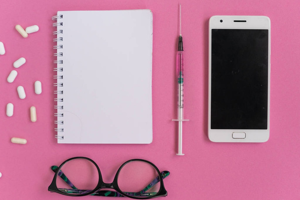 a pink background is a smartphone, a notepad, a syringe, pills and glasses - Φωτογραφία, εικόνα