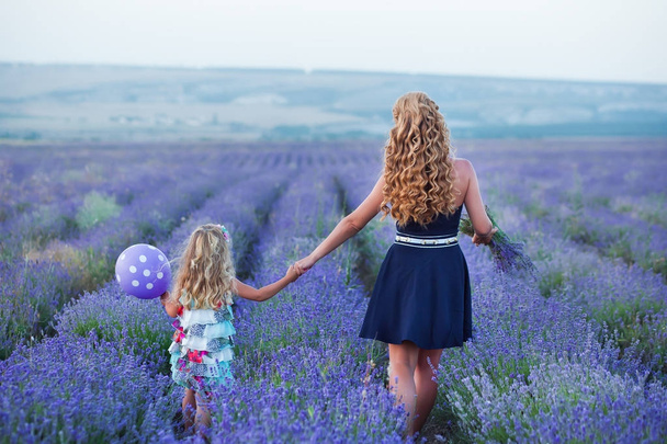 Young mother with young daughter smiling on the field of lavender .Daughter sitting on mother hands.Girl in colorful dress and mother in dark blue dress. - Фото, изображение