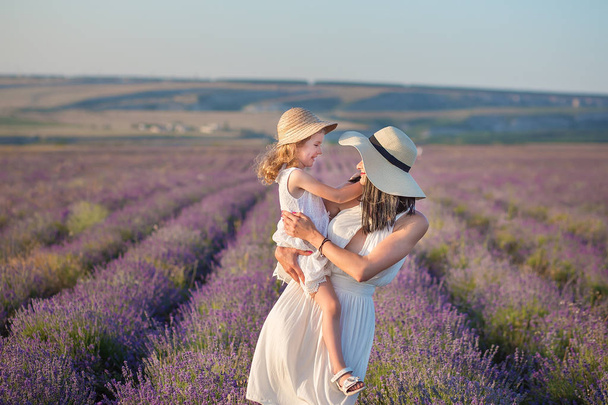Young beautiful lady mother with lovely daughter walking on the lavender field on a weekend day in wonderful dresses and hats. - Фото, изображение