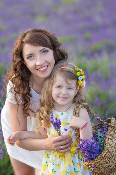 Young mother with young daughter smiling on the field of lavender .Daughter sitting on mother hands.Girl in colorful dress and mother in dark blue dress. - Photo, Image