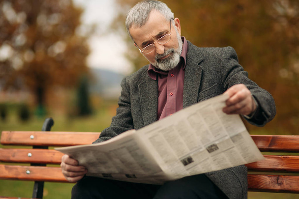 A nice grandfather with a beautiful beard in a gray jacket sits on a bench in the park and reads a newspaper - Фото, изображение