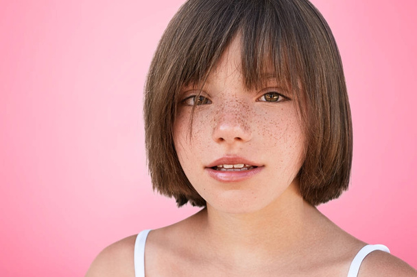 Indoor shot of confident freckled beautiful small female child with bobbed hairstyle looks at camera, glad to be photographed in studio, poses against pink background. Little kid looks at you - 写真・画像