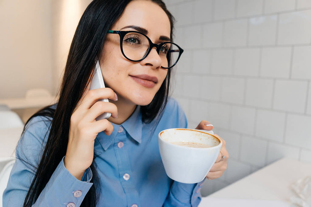 confident young girl in glasses and blue shirt talking on the phone about business, drinking coffee - Fotografie, Obrázek