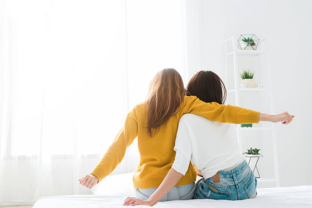 Back view of women lesbian happy couple waking up in morning, sitting on bed, stretching in cozy bedroom, looking through window. Funny women after wakeup. Lesbian couple together indoors concept. - Photo, image
