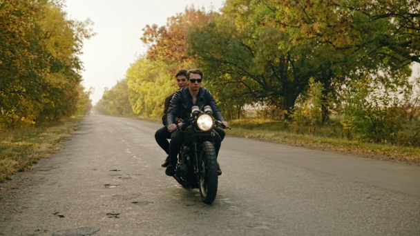Knappe man in zonnebril zitten met zijn vriendin achter het stuur van een motorfiets en het rijden op het asfaltweg in bos in de herfst - Video