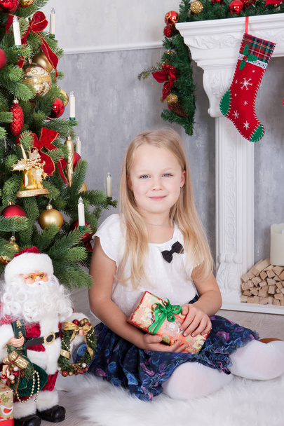 Christmas or New year celebration. Young beautiful girl in the hands holding christmas gift sitting near Christmas tree. A fireplace with christmas stocking on background - Photo, Image