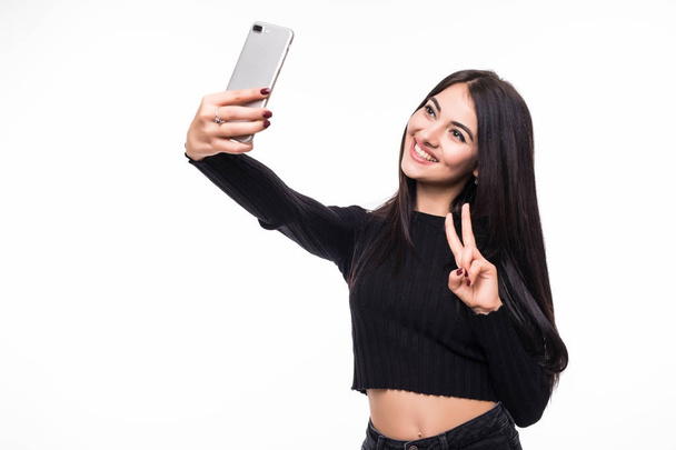 Beautiful young woman is making selfie photo with victory gesture with smartphone on white background - Zdjęcie, obraz