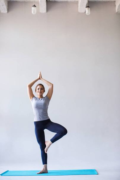 Mulher sorridente em sportswear praticando ioga, de pé no exercício Vrksasana com namaste, pose de árvore em fundo cinza
. - Foto, Imagem