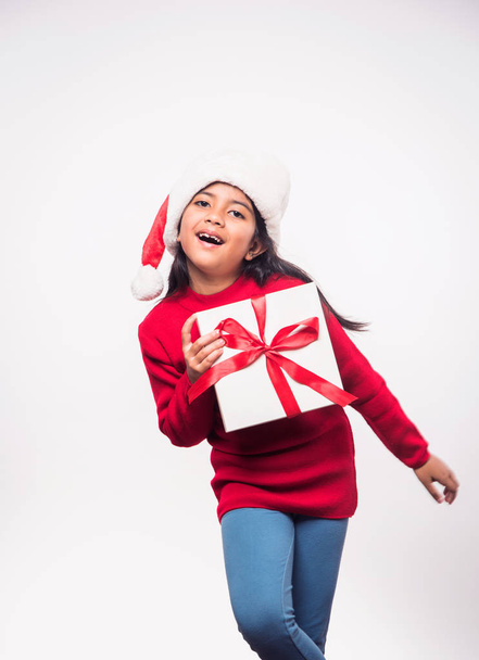 χαριτωμένο μικρό ινδικό κορίτσι φορώντας Σάντα καπέλο και γιορτάζει τα Χριστούγεννα, ενώ στέκεται απομονωμένο πάνω από λευκό φόντο - Φωτογραφία, εικόνα