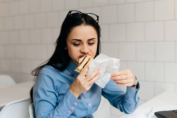 busy business girl freelancer in a blue shirt eating a sandwich in a cafe - Zdjęcie, obraz