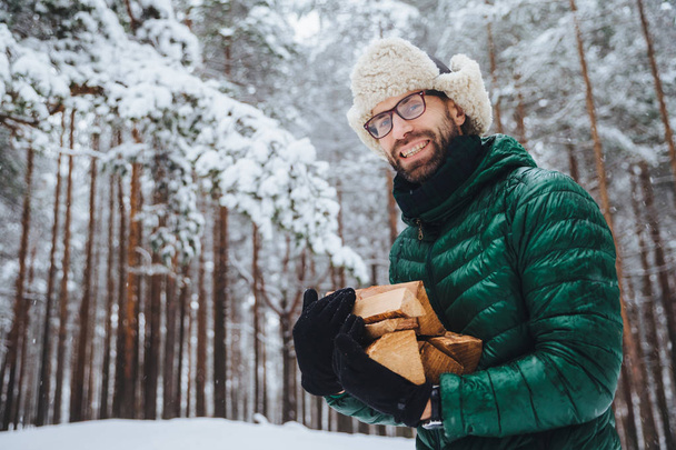 Εξωτερική πλάνο της χαμογελάει ευτυχής άνδρα με μούσι και μουστάκι που φοράει γυαλιά, anork και ζεστό καπέλο, κατέχει τα καυσόξυλα, στέκεται ενάντια δέντρα καλυμμένα με χιόνι, απολαμβάνει φρέσκο παγωμένο αέρα στο δάσος - Φωτογραφία, εικόνα