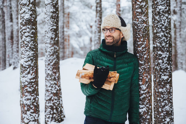Happy bearded man in green jacket, stands near tree, holds firewood, looks thoughtfully aside, has cheerful expression, stands against snowy forest background. Fashionable pensive male outdoor - Photo, Image